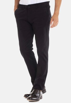 Pantalons noirs en tissu lisse, avec une coupe ajustée, des poches latérales et une fermeture à bouton. Portés avec des chaussures de ville noires.