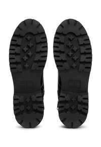 Suelas de botas negras y resistentes con profundas ranuras y "Tommy Jeans" grabado cerca del talón, vistas desde abajo sobre un fondo blanco.