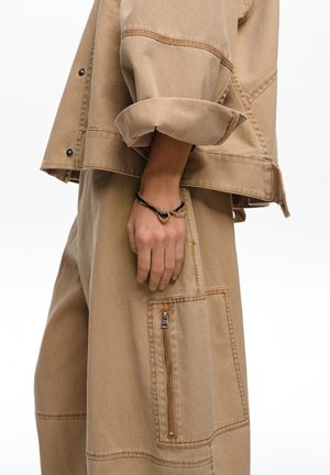 Osoba w beżowej kurtce z podwiniętymi rękawami i dopasowanych spodniach z boczną kieszenią na zamek, pokazująca na nadgarstku czarno-złotą bransoletkę z zawieszką.