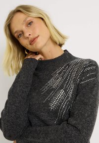Pull en tricot gris foncé avec un motif texturé d'ornements argentés sur une épaule, col montant et manches longues.