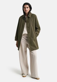 Trench coat sin forro en verde oliva con cuello alzado y botones al frente. Combinado con pantalones anchos color crema y zapatillas, presenta un ajuste relajado.