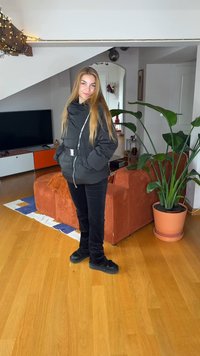 Μαύρο μπουφάν τύπου puffer με ζώνη και κλείσιμο με φερμουάρ, συνδυασμένο με μαύρο παντελόνι και σκούρα παπούτσια, στέκεται σε ένα δωμάτιο με φυτά και έπιπλα.