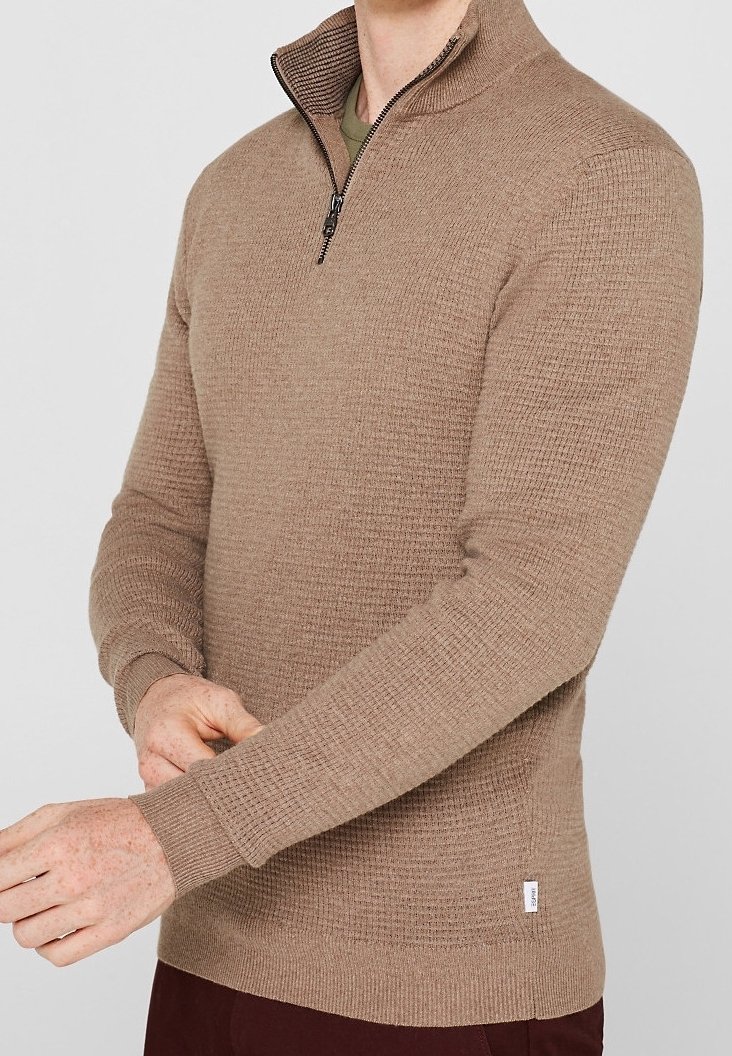 Homme ajustant la manche d'un pull beige texturé à fermeture éclair quart de zip par-dessus une chemise verte, portant un pantalon marron foncé sur fond uni.