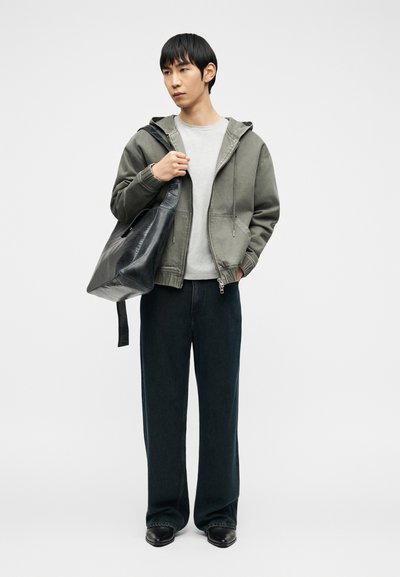 Νεαρός άνδρας με γκρι φούτερ με φερμουάρ, ανοιχτό γκρι μπλουζάκι, φαρδιά μαύρα παντελόνια, μαύρα παπούτσια, κρατώντας μια μεγάλη μαύρη δερμάτινη τσάντα ώμου.