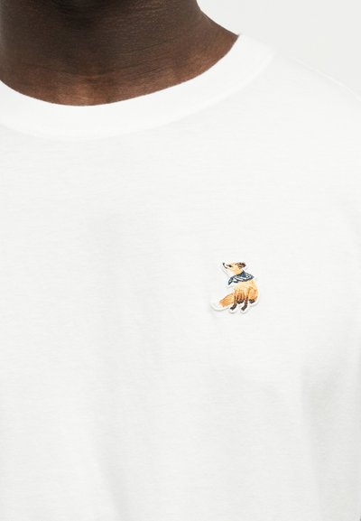 Λευκό μπλουζάκι με στρογγυλή λαιμόκοψη και μικρή κεντημένη αλεπού με μπλε κασκόλ στο αριστερό στήθος, φορεμένο από άτομο με σκουρόχρωμο δέρμα.