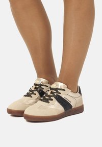 Scarpe da ginnastica beige con tomaia texturizzata, dettagli neri, accessori dorati e lacci a fantasia. Suola in gomma. Design con collo piatto.