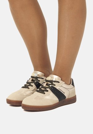Beige Sport Schuhe mit einer strukturierten Oberseite, schwarzen Akzenten, goldenen Beschlägen und gemusterten Schnürsenkeln. Gummisohle. Flaches Fußgewölbedesign.