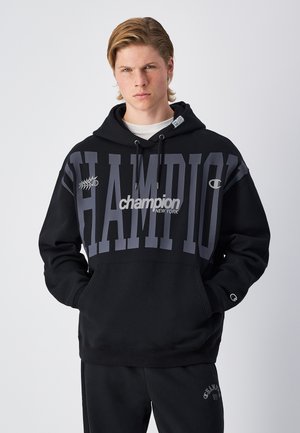 Black hoodie with large "CHAMPION" logo in gray across the front, adjustable drawstring hood, and front pocket. Soft fabric texture.