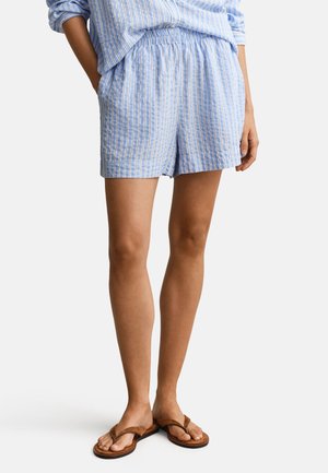 Femme portant un short à rayures verticales bleu clair et blanc assorti à un haut à manches longues et des tongs marron, debout les mains dans les poches.