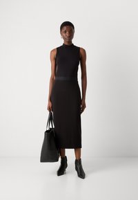 Robe noire sans manches avec un col montant et une coupe ajustée. Associée à des bottines et portant un sac fourre-tout noir. Texture lisse.