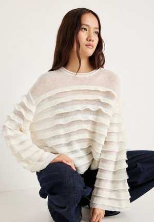 White striped knit sweater with a sheer, textured design, featuring horizontal layered accents and wide sleeves. Worn with dark denim pants.
