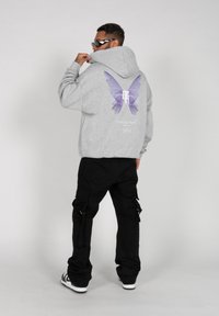 Sudadera gris con gráfico de mariposa morada en la parte posterior, material de algodón, acompañada de pantalones cargo negros y gafas de sol elegantes.