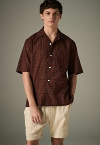 Chemise marron à manches courtes avec des motifs découpés, dotée d'une patte de boutonnage et de boutons bleu clair, associée à un short crème texturé.