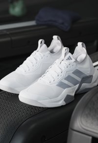Coppia di scarpe sportive bianche con accenti grigi posizionate su una superficie nera testurizzata di un tapis roulant interno.