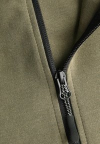 Tessuto verde oliva con fine trama, caratterizzato da una cerniera nera lungo la cucitura. Elementi notevoli includono il tirazip con impugnatura testurizzata.