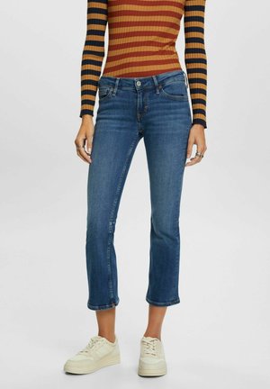 Femme portant un jean court bleu, des baskets blanches et un haut côtelé à manches longues avec des rayures horizontales orange et noires.