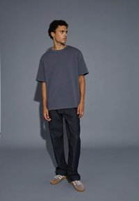 Junger Mann steht, trägt ein locker sitzendes graues T-Shirt, dunkle Jeans und beige Turnschuhe mit weißen Streifen vor einem schlichten grauen Hintergrund.