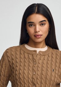 Ung kvinna med långt mörkt hår, som bär guldfärgade hoopsörhängen och en brun kabelstickad cardigan med knappar över en ljus topp.