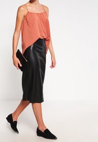 Top in raso ruggine con spalline sottili, abbinato a una gonna midi in pelle nera. Presenti anche delle scarpe nere senza lacci e una pochette.