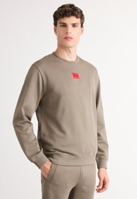 Felpa color khaki con collo a giro e polsini a coste, dotata di un patch rettangolare rosso con logo sul petto. Tessuto morbido e texturizzato.