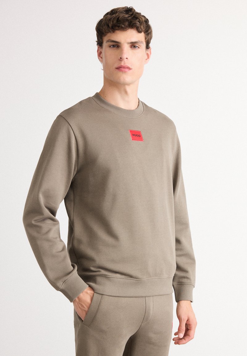 Felpa color khaki con collo a giro e polsini a coste, dotata di un patch rettangolare rosso con logo sul petto. Tessuto morbido e texturizzato.