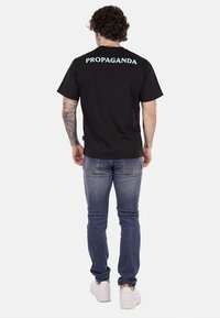 T-shirt di cotone nera a maniche corte con la scritta "PROPAGANDA" in azzurro sul retro. Il modello indossa jeans blu e sneakers bianche.