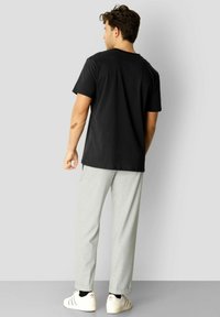 Svart bomull T-skjorte med rund hals, kombinert med lysegrå bukser og hvite joggesko med røde detaljer. Sett fra baksiden.