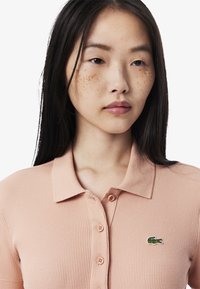 Vaaleanpunainen ribbitoppi, jossa on kaulus ja neljä nappia, sekä vihreä brodeerattu krokotiili-logo vasemmassa rinnassa.