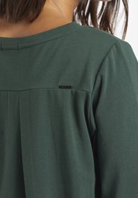 Maglietta a maniche corte verde scuro realizzata in tessuto morbido, con dettaglio a pieghe sulla parte superiore della schiena e un piccolo accento con logo nero.