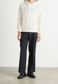Beige hoodie med framficka och dragsnören, kombinerad med svarta raka byxor och svarta läderskor på en neutral bakgrund.