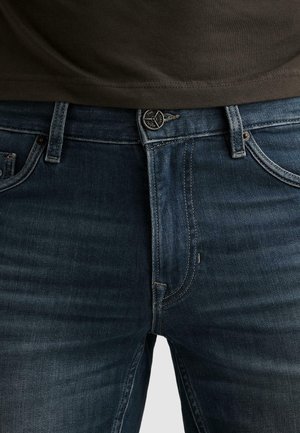 Detail tmavě modrých džín s předním knoflíkem a zipem, nošených s tmavě hnědou košilí zasunutou mírně nad pasovou linií.