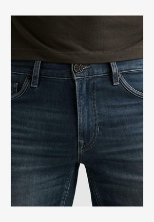 Gros plan sur un jean en denim bleu foncé avec bouton et fermeture éclair à l'avant, porté avec une chemise marron foncé rentrée légèrement au-dessus de la ceinture.