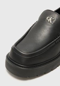 Scarpa slip-on in pelle nera con suola piatta, dettagli di cucitura in rilievo e logo "CK" in argento sulla parte anteriore superiore.