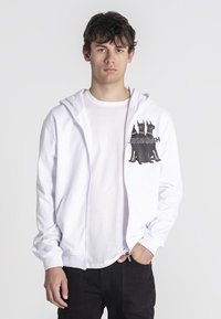 Biela mikina na zips s grafickou potlačou dvoch čiernych psov na ľavej strane hrudníka, nosená cez jednoduché biele tričko a čierne nohavice.