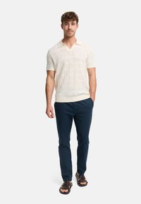 Homme portant un polo tricoté à manches courtes crème, un pantalon bleu marine et des sandales marron foncé, debout devant un fond uni.