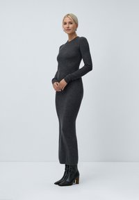 Dlouhé, přiléhavé šedé pletené šaty s dlouhými rukávy, kulatým výstřihem a hladkou texturou, doplněné o černé kotníkové boty na podpatku.