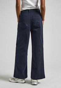 Plaši, tumši zila kokvilnas bikses ar divām aizmugurējām kabatām, ar viļņotu šuvju detaļām un brīvu piegriezumu, saskaņotas ar gaišām kurpēm.