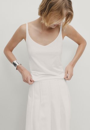 Femme aux cheveux blonds courts ajustant son haut sans manches blanc, portant une jupe blanche et un bracelet en argent à son poignet gauche.