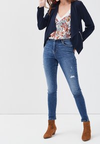 Femme portant un blazer bleu marine, un haut à fleurs, un jean skinny taille haute et des bottines en daim marron, se tenant avec une main près du visage.