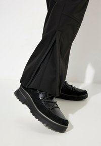 Black footwear with a textured upper, lace detail, and a thick, rugged sole. Pants feature side zippers and a smooth fabric finish.