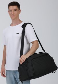 Borsa da viaggio nera realizzata in materiale resistente, dotata di una tracolla, maniglie superiori e più scomparti con zip per la conservazione.