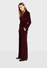 Burgundska baršunasta odijela s krojenim blejzerom s izrezanim reverima, dva dugmeta i širokim hlačama. Crne ravne cipele upotpunjuju izgled.