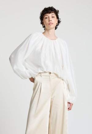 Blouse blanche volumineuse à manches longues avec un col rond, associée à un pantalon crème taille haute avec plis et ceinture.