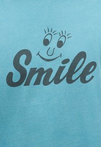 Μπλε ύφασμα με τη λέξη «Smile» σε μαύρη γραφή, συνοδευόμενη από ένα χαμογελαστό πρόσωπο με μάτια και βλεφαρίδες πάνω από το κείμενο.