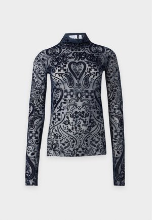 Femme portant un haut noir transparent à manches longues avec un motif et un jean gris ample avec de grandes pièces marron aux genoux, debout devant un fond uni.