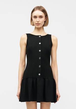 Γυναίκα που φορά αμάνικο μαύρο φόρεμα με κουμπιά μπροστά και φαλτσαριστό τελείωμα, στέκεται μπροστά σε απλό φωτεινό φόντο.