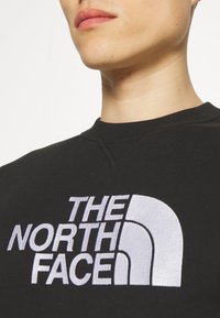 Sudadera negra con cuello redondo, que presenta un gran logotipo bordado en blanco "THE NORTH FACE". Tejido suave con una textura lisa.