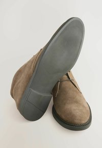 Brune ruskind ankelstøvler med rund tå, flad gummisål og minimal snørebåndsdetalje. Glat tekstur med stikninger omkring sålen.