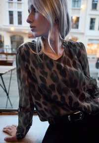 Transparente Bluse mit Leopardenmuster in Dunkelgrün und Schwarz, mit weitem Ausschnitt und langen, gepufften Ärmeln; kombiniert mit dunklen Jeans und einem Gürtel.