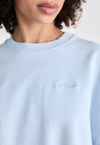 Lyseblå hettegenser laget av myk stoff, med ribbestrikket rund hals og brodert logo på brystet i en subtil, matchende farge.